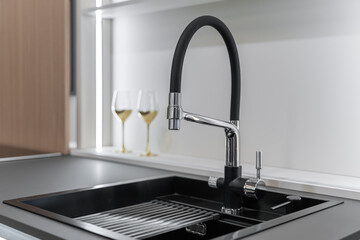black kitchen sink and black faucet close-up. Black tabletop and white backsplash. Wine glasses....