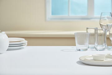 Clean glass cups, bowls and plates are arranged on the white kitchen table with bright light from the window, evoking a fresh and tidy atmosphere after cleaning.