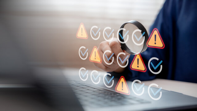 Hand using magnifying glass to inspect warning and check icons on laptop screen, representing system monitoring, debugging, error detection, and cybersecurity analysis.