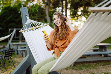 Stylish woman with wireless headphones and phone relaxing lying in hammock in sunny park. Beautiful...