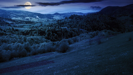 vast rural landscape in mountain at night. rolling hills with forest and pasture in full moon light. starry sky with dark clouds
