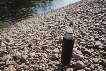 A Stainless Steel Thermos Bottle rests gracefully on the Pebbly Riverbank, inviting you to embark on an adventure filled with outdoor activities like hiking, camping, and serene picnics by the beach