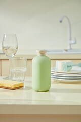 Green bottle and dish sponge beside crystal glasses on a beige surface, blending utility and elegance in a clean kitchen layout.