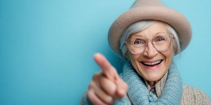 Joyful senior female in a stylish hat and eyewear, playfully posing against a pastel backdrop.