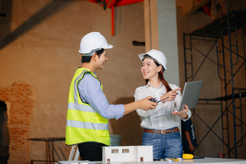 Asian engineers discuss floor plan using laptop and building model at active construction site. Collaboration, safety