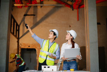 Asian engineers discuss floor plan using laptop and building model at active construction site....