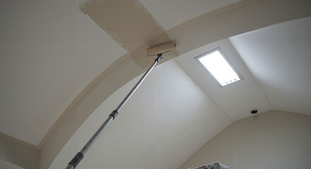 Painting a high interior ceiling with a paint roller on an extension pole. Home improvement and DIY renovation project, applying a fresh coat of beige paint to the surface.
