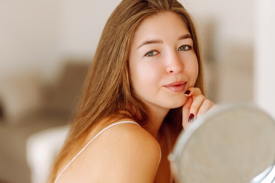Portrait of a cute woman with a mirror looking at her reflection. Young woman enjoying the sun in a cozy room. Dermatology and beauty concept.