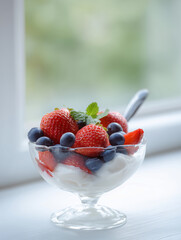 Glass bowl of mixed berries (strawberry, blueberry) with yogurt, natural window lighting, healthy breakfast style