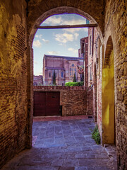 The church of Basilica Cateriniana di San Domenico in Siena, Tuscany, Italy, emerges beautifully through an old arched alley, framed by historic stone walls and bathed in soft natural light.