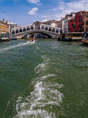 View of the Rialto Bridge in Venice with boats crossing the troubled waters of the Grand Canal, and a blue sky with sparse clouds overhead.