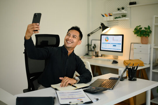 A cheerful businessman sitting at his desk, smiling and holding a smartphone during a video call in a modern office - Powered by Adobe