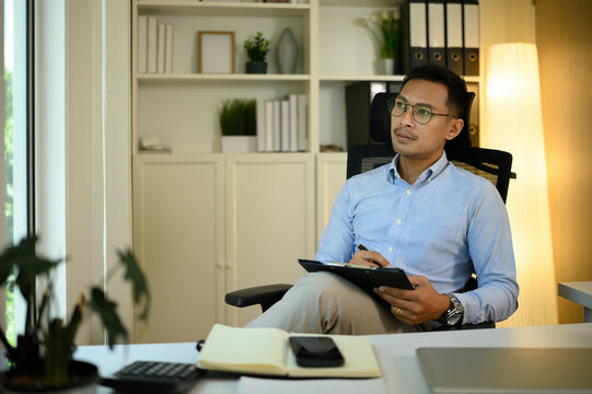 A mature Asian businessman sits thoughtfully with a clipboard in hand, contemplating ideas in a modern office