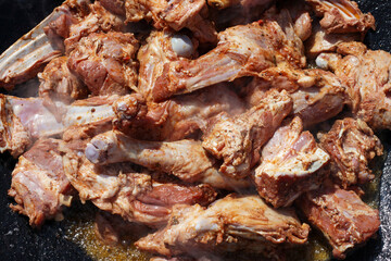 Close-up of seasoned meat cuts with bones, sizzling in a large outdoor cooking pan, creating a rich aroma and appealing texture