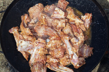 Overhead view of savory meat cuts with bones, sizzling and browning in a large outdoor cooking pan, creating a delicious aroma