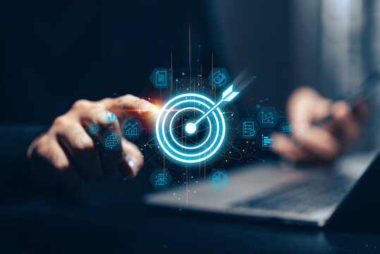 Businessman holding a glowing blue target with arrow in his palm above a laptop, surrounded by holographic business icons representing goals, analytics, strategy, and online presence. This conceptual.