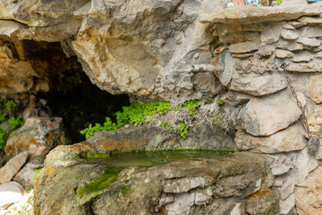 Close-up shows ancient natural spring with hand-forged metal spout trickling water into shallow rock basin, surrounded by lush green moss and creeping stone textures