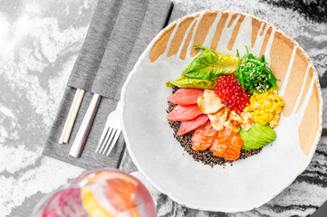 A vibrant poke bowl with salmon, tuna, avocado, corn, seaweed, lettuce, and red fish roe on black quinoa, served on a marble table with utensils and a drink