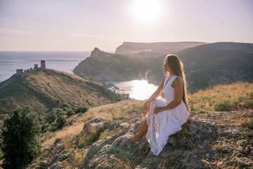 Naklejka premium A woman in a white dress sits on a rock overlooking a body of water. The scene is serene and peaceful, with the woman enjoying the view and the calmness of the surroundings.
