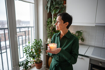 Joyful calm mature woman enjoying candlelight at home, self care ritual, time with yourself. Stress relief, emotional wellbeing, home decoration, mindfulness breathing, natural scent therapy