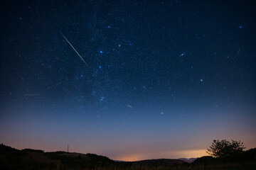 Milky Way stars with meteor shower trails and countryside silhouettes.