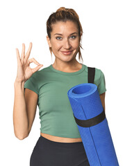 Blonde athlete holding a yoga mat cheerful and confident showing ok gesture.
