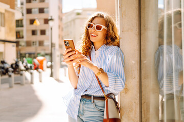 Stylish young woman wearing sunglasses and holding a phone in hands, enjoying walk in the old town....
