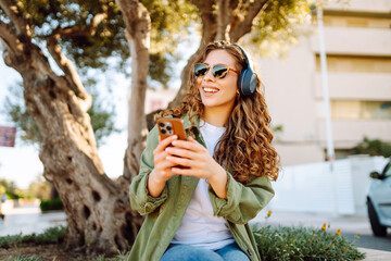 Stylish woman in sunglasses with phone and wireless headphones enjoying weather in sunny park....