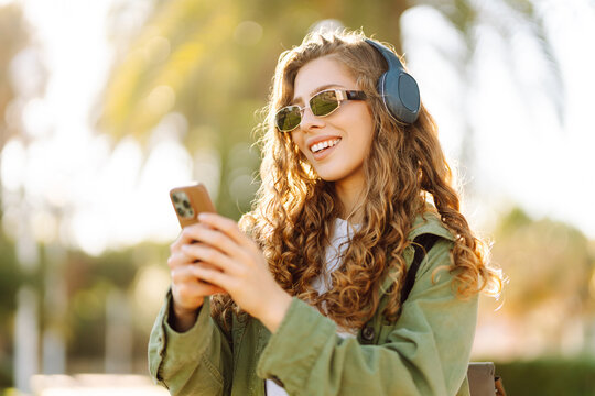 Stylish woman in sunglasses with phone and wireless headphones enjoying weather in sunny park. Beautiful tourist walking along embankment outdoors. Concept of technology and enjoyment.