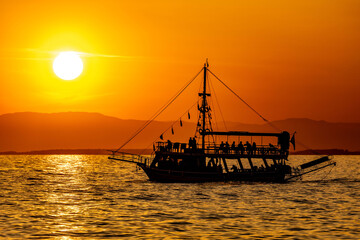 Obraz premium Silhouette of a tourist boat sailing at sunset in Side, Turkey. Golden sun paints the Mediterranean sea with vibrant colors. Majestic mountain backdrop.