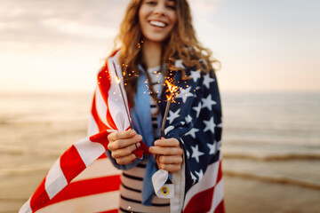 Young woman wrapped in American flag holding sparklers on sunset beach. Beautiful woman enjoying...