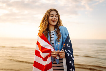 Young woman wrapped in American flag holding sparklers on sunset beach. Beautiful woman enjoying...