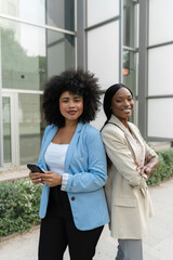 Vertical image of Two multiracial businesswomen standing outside office building smiling at camera
