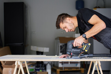 Worker in workwear using handheld cordless electric jigsaw to cut wooden board in garage. Skilled...