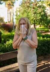 Senior woman blowing nose with tissue in park suffering from allergies