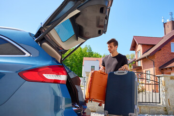 Man holding orange and blue suitcases near parked car in front of suburban brick house. Loading...