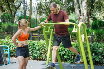 Man exercising on outdoor fitness equipment in a park, assisted by a female trainer.  They are using a leg press machine for a lower body workout. Healthy lifestyle and fitness concept.