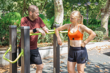 Young man and woman resting after a workout at an outdoor gym. They are leaning against a horizontal bar in a park, possibly discussing their training.  The image conveys a sense of fitness and