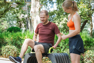 Man doing outdoor workout in park with female trainer.  He is using outdoor gym equipment.  Fitness, exercising, healthy lifestyle concept.