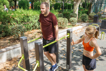 Man doing parallel bar dips outdoor in a park.  A woman assists him with his workout.  Calisthenics training and fitness in a public space.