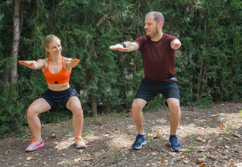 Active couple performs squats outdoors in a park.  They are wearing sportswear and fitness shoes. Healthy lifestyle and fitness concept.