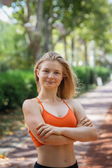 Confident young blonde woman in sportswear stands with arms crossed in a park.  She's wearing an orange sports bra and black shorts. Healthy lifestyle, fitness, and well-being concept.