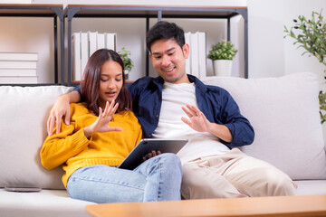 Obraz premium Happy asian couple video call on tablet sitting on sofa in living room at home, smiling and waving during online communication with relax, couple watching tablet, lifestyle and interaction.