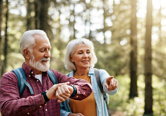 Active senior couple hiking in forest with backpacks, man checking smartwatch while woman points ahead, enjoying nature together