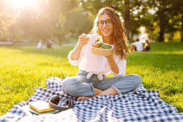 Happy woman in glasses eating salad on a picnic mat. Young woman enjoying a healthy bowl and...