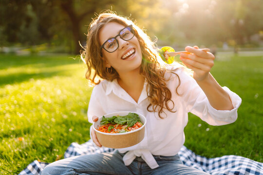 Happy woman in glasses eating salad on a picnic mat. Young woman enjoying a healthy bowl and basking in the sun in a sunny park. Concept of relaxation, enjoyment.