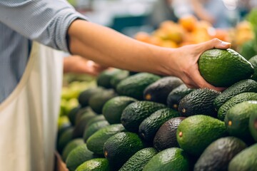 A person selecting a ripe avocado from a pile at a market or grocery store with a blurred background.