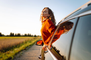 Cheerful woman leaning out of her car window feeling free. Young woman traveling by car enjoying...