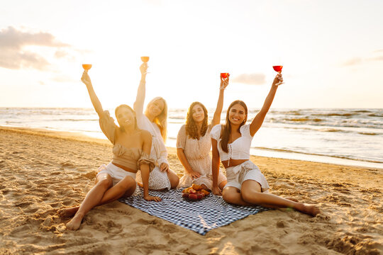 Cheerful female friends spending time together on picnic on sunset beach. Four young women with glasses in hands enjoying seascape. Friendship and fun concept. - Powered by Adobe