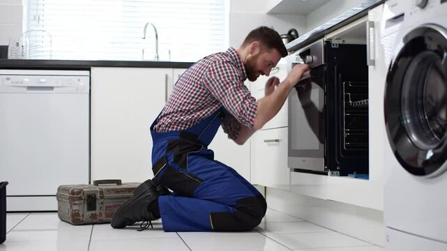 Handyman in Blue Overalls Repairs a Black Oven in a Modern White Kitchen with Toolbox and Washing Machine Under Bright Daylight